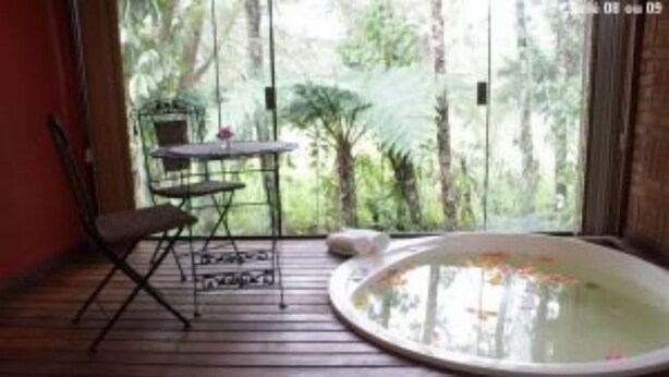 A room with a wooden floor, a round hot tub, a small table with a chair, and a view of greenery through a glass door. - Pousada no Caminho do Marimbondo (Itatiaia)
