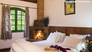 A cozy room with a bed, a fireplace, a window with curtains, and a painting on the wall. - Pousada no Caminho do Marimbondo (Itatiaia)