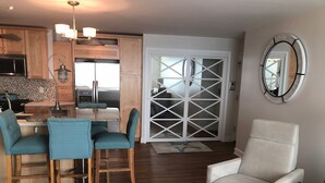 A modern kitchen with wooden cabinets, a dining area with teal chairs, and a white armchair. - BEACHFRONT LAKE FACING Lake Michigan 1BR - private beach/heated pool/sundeck! (South Haven)