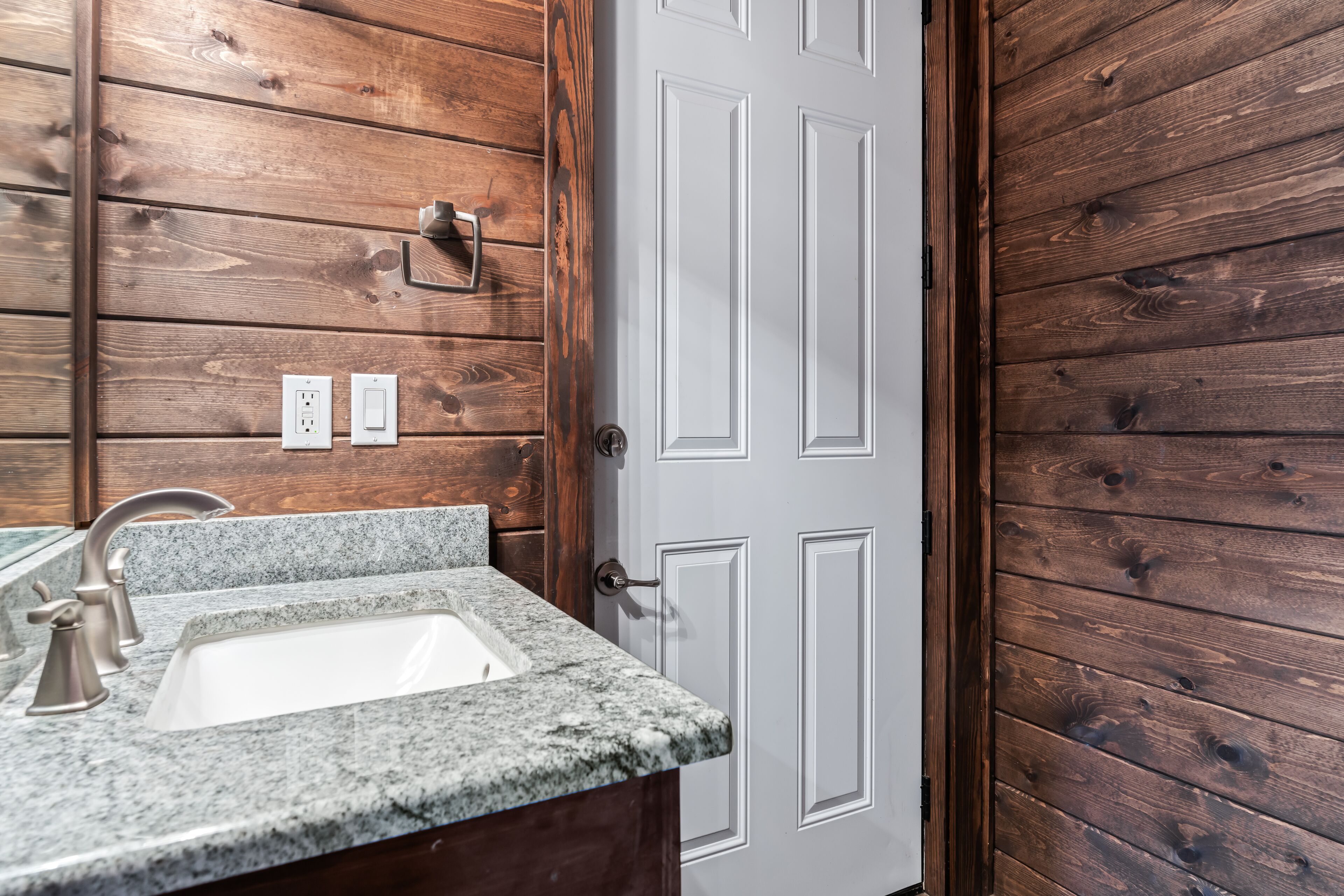 Un baño con lavamanos de encimera de granito, paneles de madera y una puerta blanca.