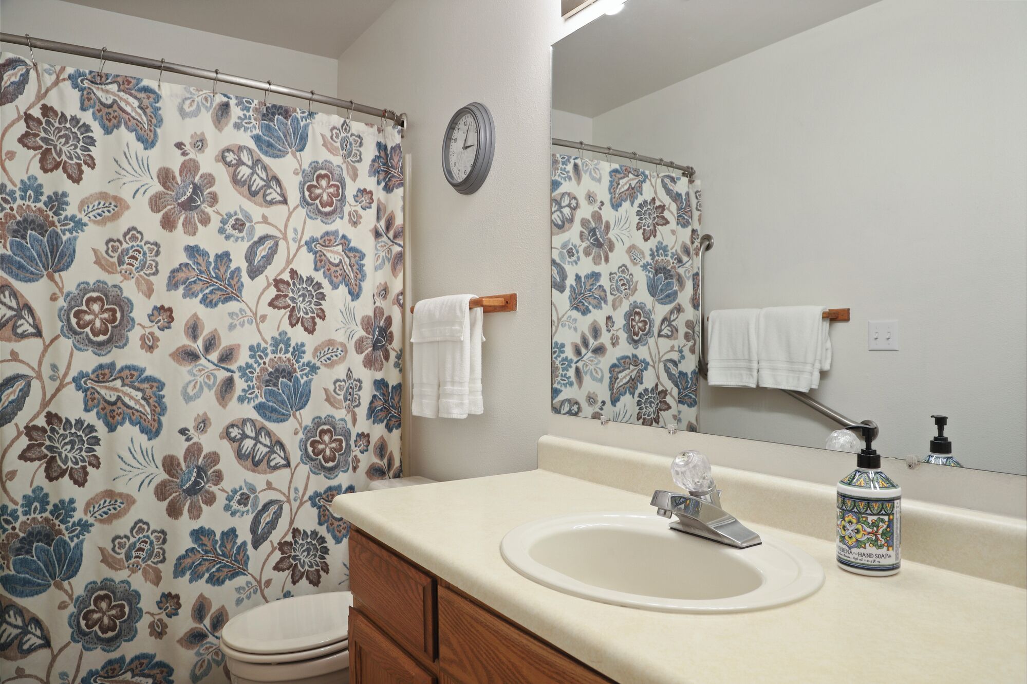 Un baño con cortina de ducha estampada con flores, un mueble de madera, un lavabo con dispensador de jabón y un espejo.