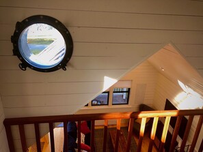 Uitzicht vanuit een raam in een kamer met houten wanden, een houten trap en een klein raam dat uitkijkt op een weg en groen.