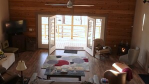 L’intérieur chaleureux d’un chalet, avec un plafond haut, des murs en bois, une cheminée centrale et une grande fenêtre donnant sur une terrasse.