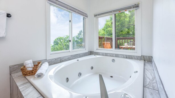 A modern bathroom with a large bathtub, two windows, a wooden deck visible through the windows, and a marble countertop.