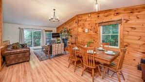 A wooden cabin interior with a dining table, chairs, a sofa, and a television.