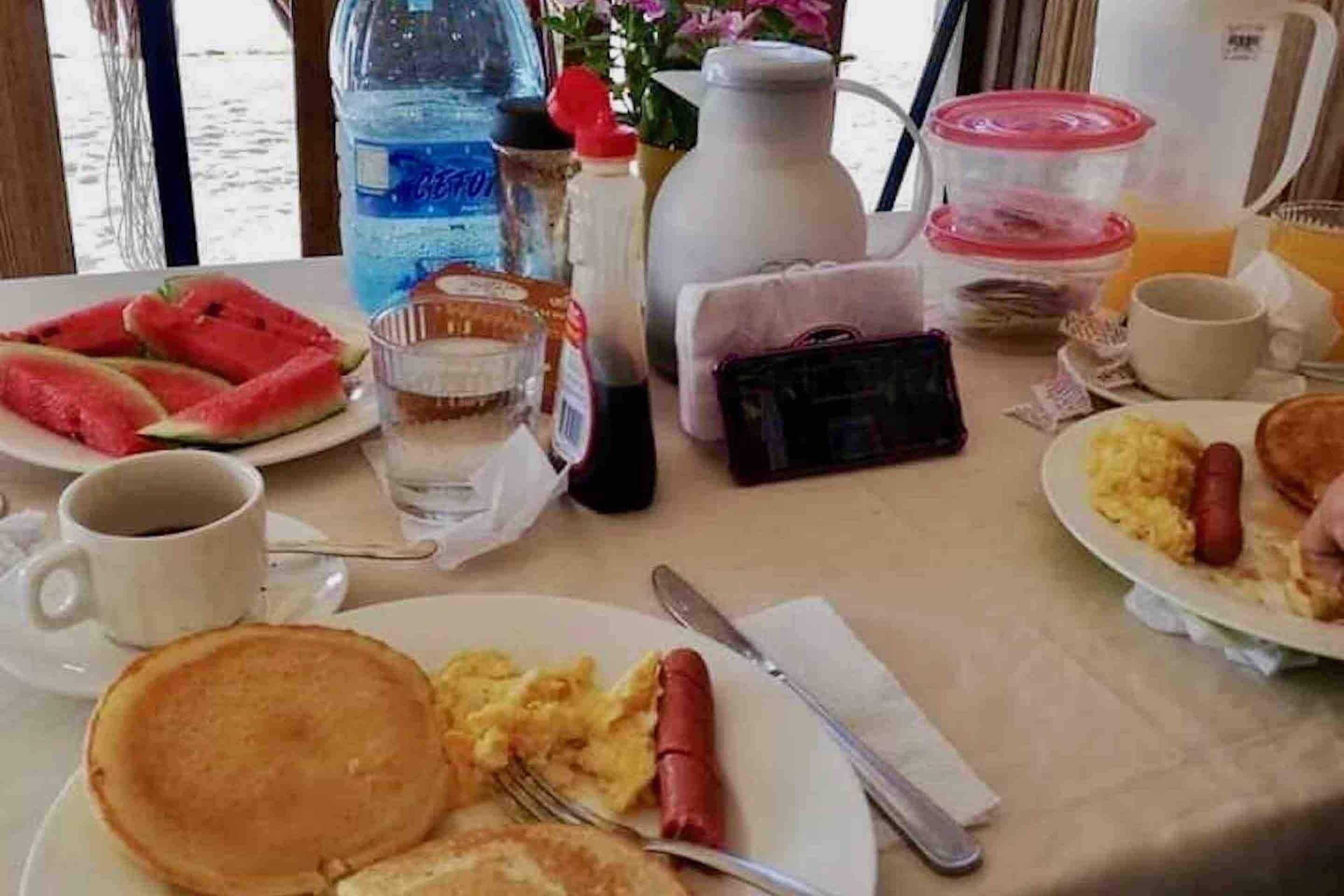 Una mesa de desayuno con platos de tortitas, huevos revueltos y un hot dog, junto con una botella de agua, una taza de café y una jarra de zumo de naranja.
