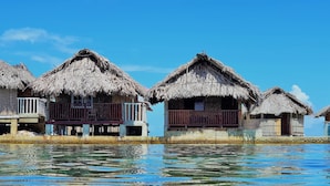 Bungalows sobre el agua con techos de paja y barandillas de madera, unidos por una pasarela de hormigón sobre aguas cristalinas.