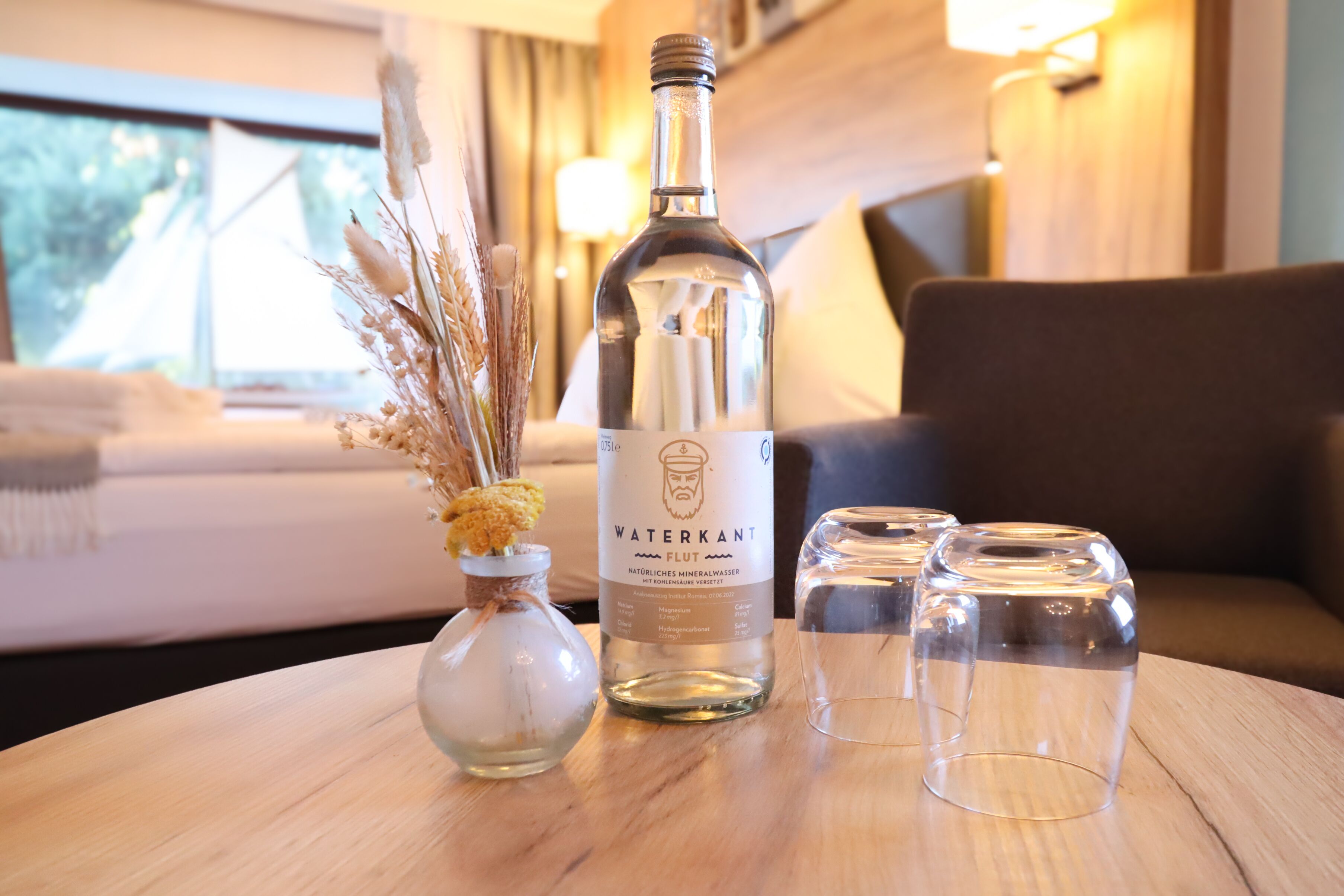 Una ampolla d'aigua mineral Waterkant, un petit vas amb flors seques i dues copes de cristall sobre una taula de fusta en una habitació d'hotel.