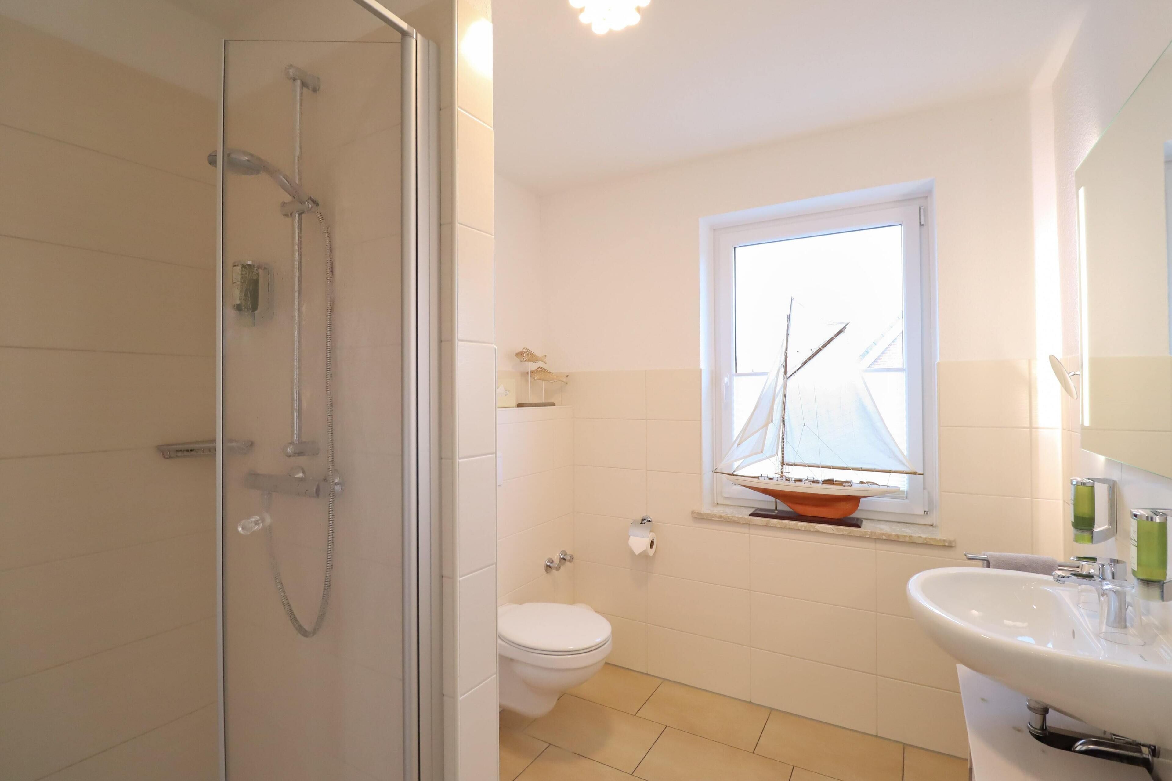 A bathroom with a glass-enclosed shower, a toilet, a sink, and a window with a sailboat decoration.