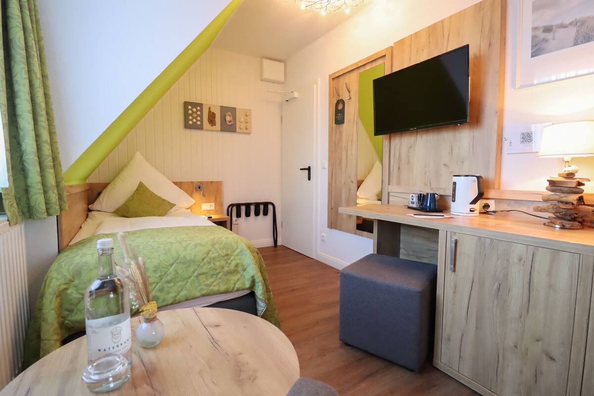 Une chambre d’hôtel compacte comprenant un lit, une petite table avec une bouteille et un verre, un téléviseur fixé au mur et un coin cuisine.