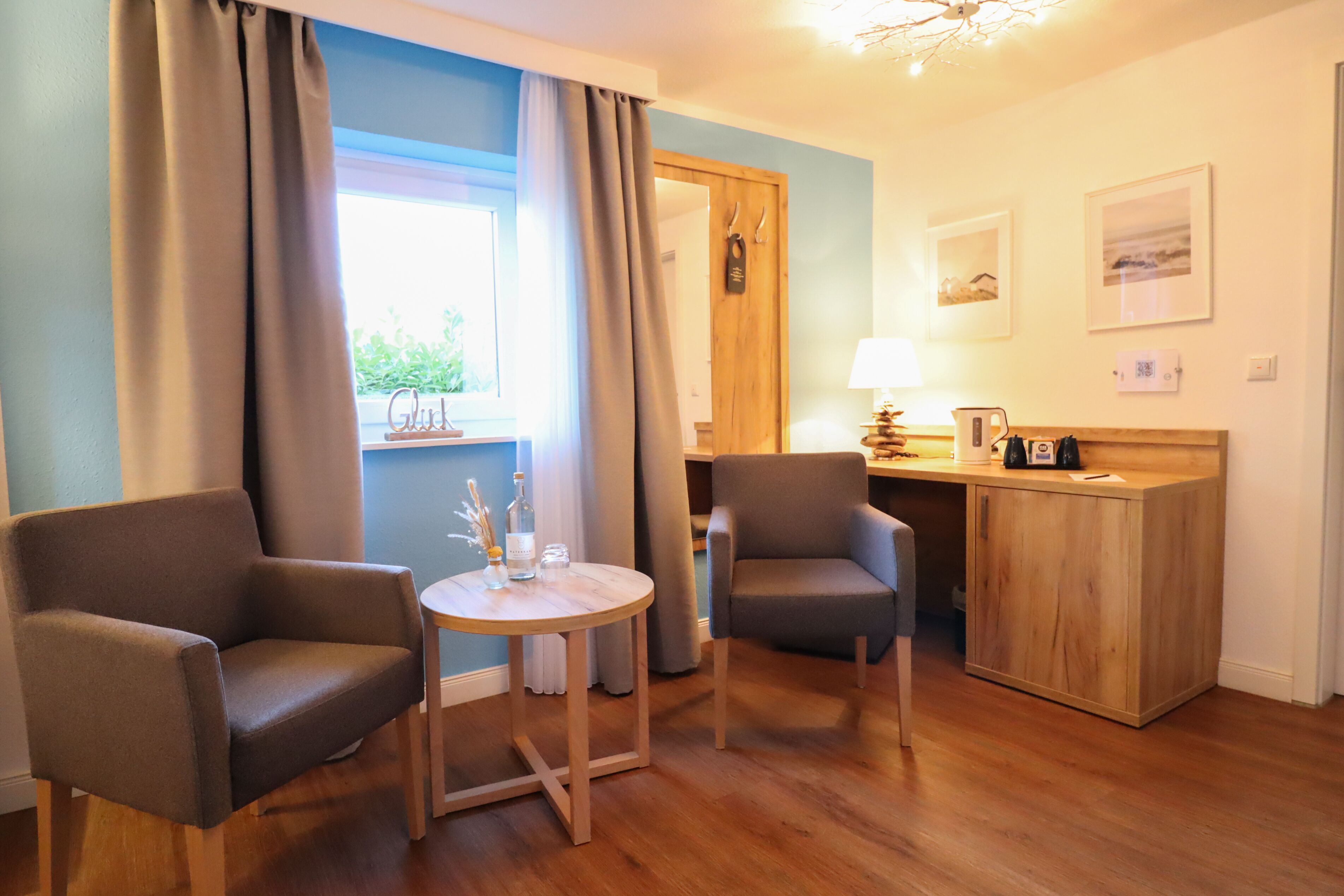 Ein Zimmer mit zwei Sesseln, einem kleinen runden Tisch, einem Holzbüro und einem Fenster mit Vorhängen.