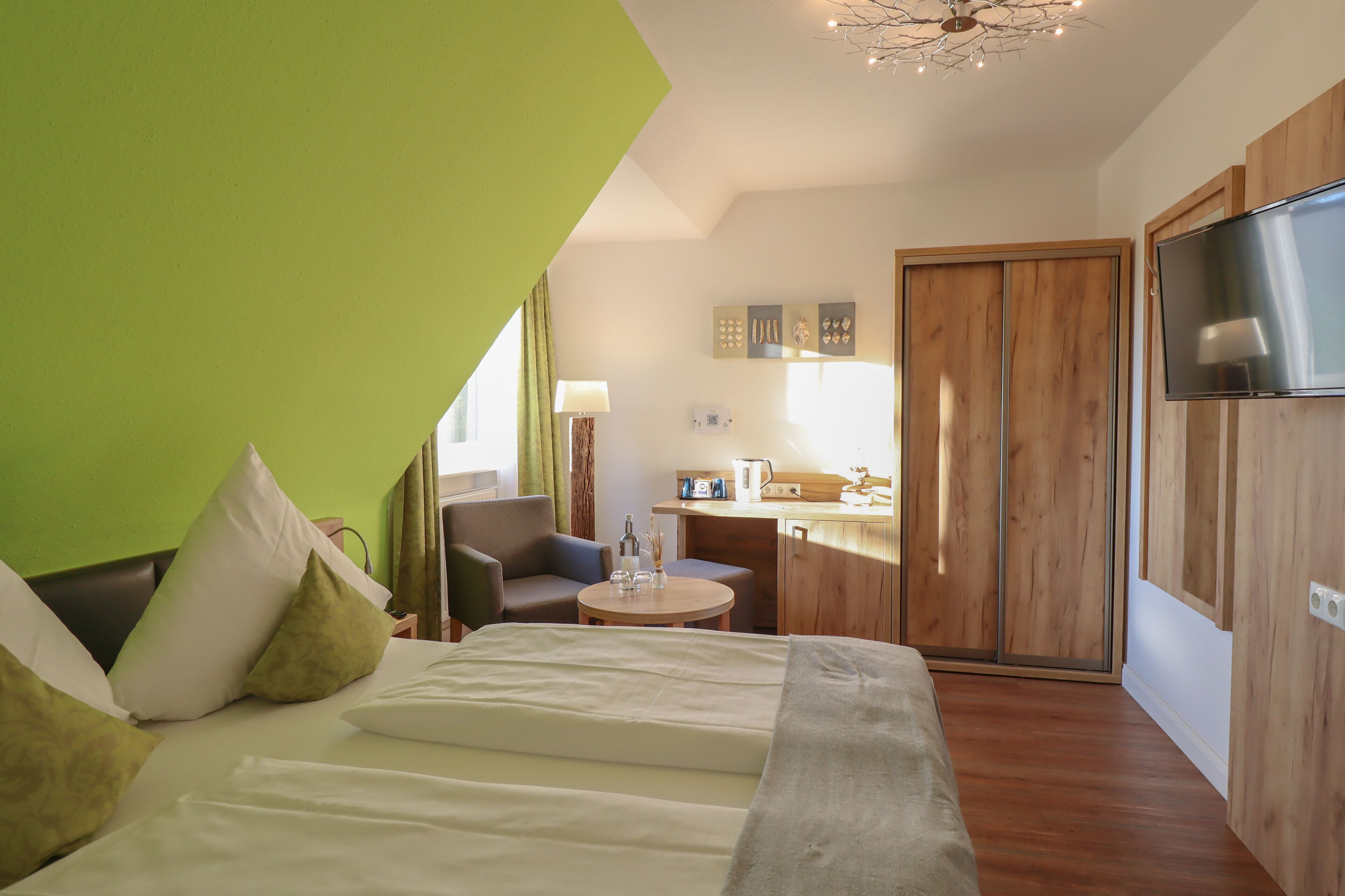 A modern hotel room with a bed, a small table, a chair, a kitchenette, and a TV.