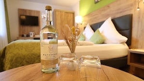 A hotel room with a bed, a TV mounted on the wall, a bottle of water, and a vase with dried flowers on a wooden table.