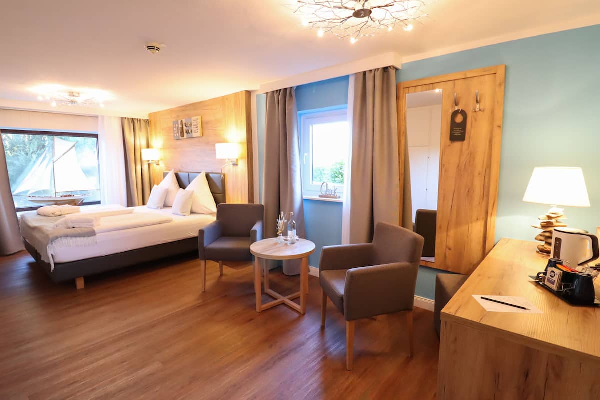 Une chambre d’hôtel comprenant un lit, deux fauteuils, une petite table ronde, un bureau avec une lampe et une fenêtre avec des rideaux.