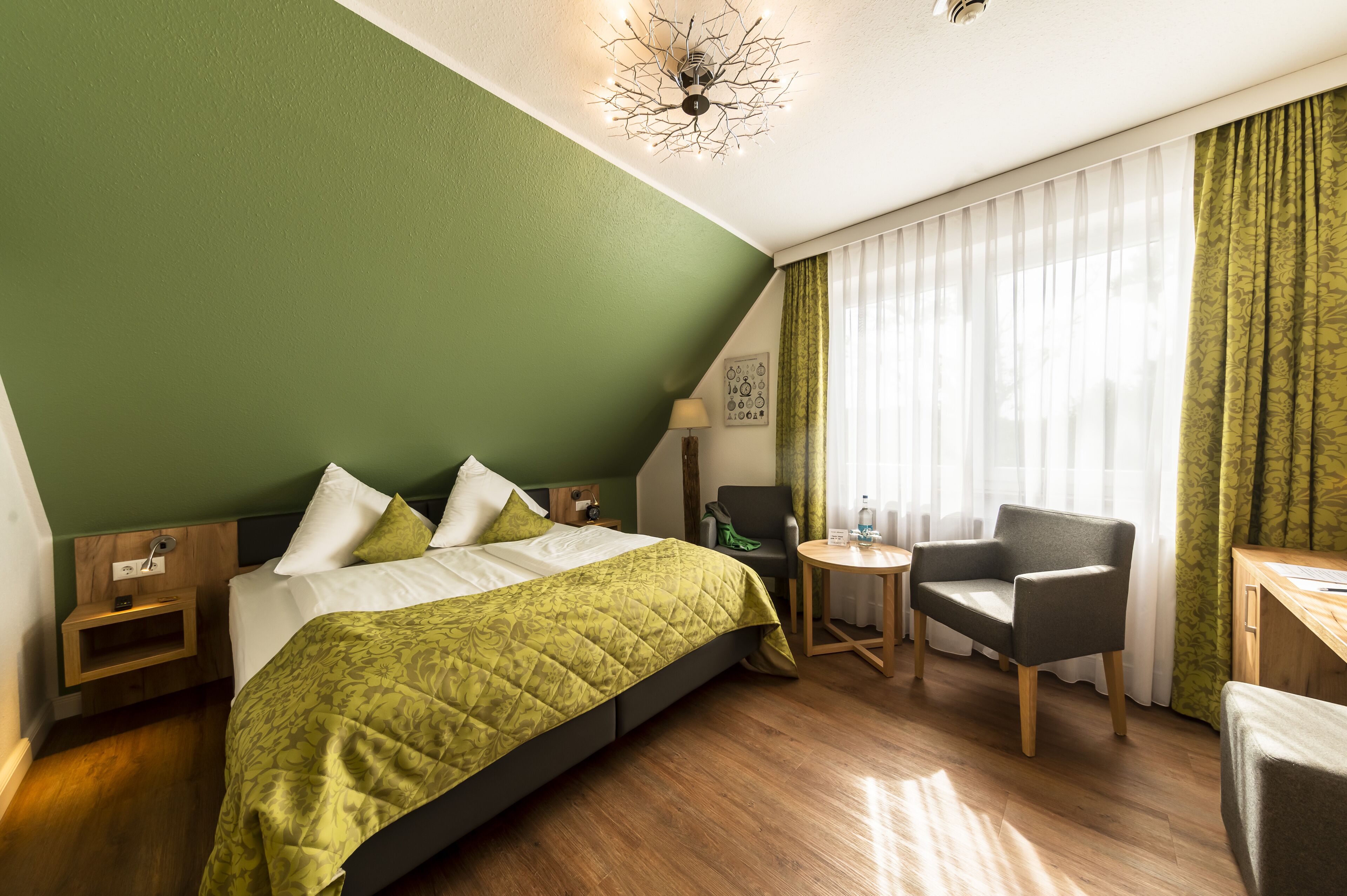 A hotel room with a green wall, a bed with a green quilt, a wooden nightstand, a chair, a small table with a lamp, and a desk with a chair.