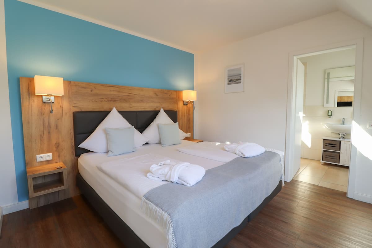 Une chambre d’hôtel avec un grand lit, une tête de lit en bois, des lampes de chevet, une petite table de chevet et une salle de bain visible à travers une porte ouverte.
