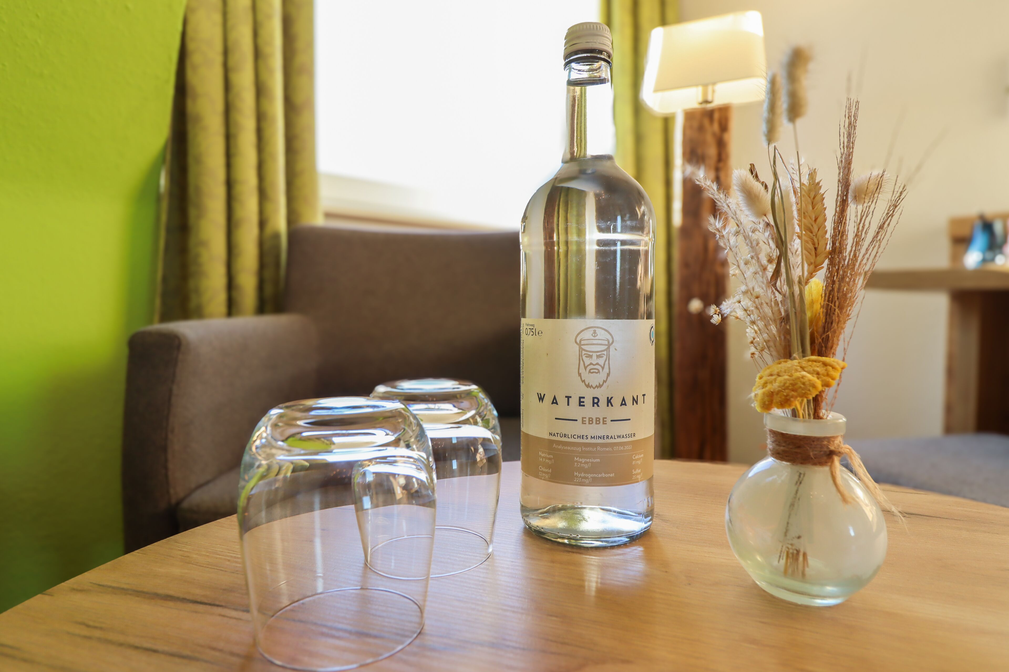 Una botella de agua mineral Waterkant Ebbe, un jarrón con plantas secas y tres vasos vacíos sobre una mesa de madera en una habitación con pared verde y una lámpara al fondo.