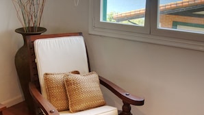 Une chaise en bois avec un coussin blanc et un oreiller tissé, placée près d’une fenêtre donnant sur la mer.