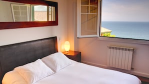 Un lit avec du linge de maison blanc, une table de chevet avec une lampe, une fenêtre donnant sur la mer et un radiateur.