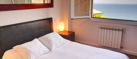 A bed with white linens, a nightstand with a lamp, a window with a view of the sea, and a radiator.