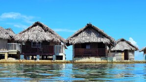 Une rangée de bungalows sur pilotis aux toits de chaume, dotés de balcons et reliés par une passerelle en bois.