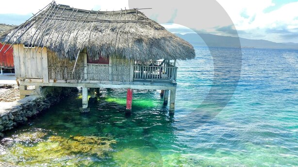 Un bungalow sur pilotis avec toit de chaume, une terrasse en bois, une eau d’un bleu cristallin et une chaîne de montagnes qui se dessine à l’horizon.