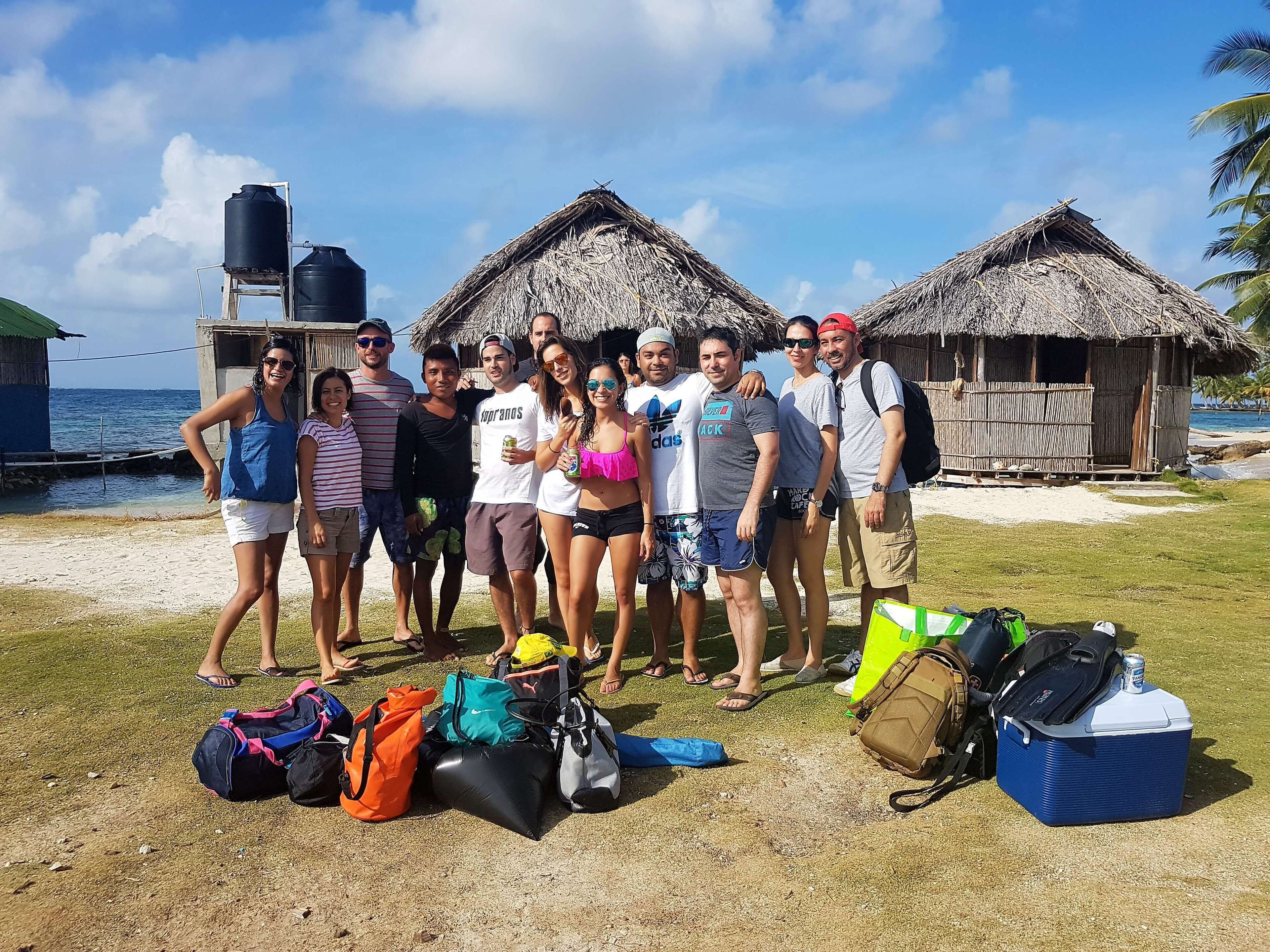 A group of people posing in front of thatched-roof huts by the sea, with luggage and a cooler on the ground.