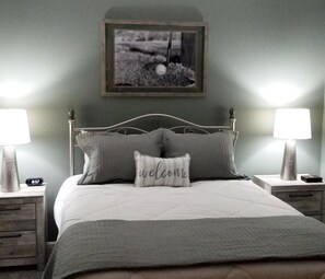 A bedroom with a bed, two nightstands, a framed picture on the wall, and a pillow with "welcome" written on it.
