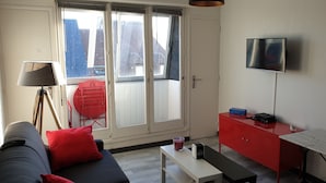 A modern living room with a sofa, a red cabinet, a TV, and a window with a view of buildings. - Apartment F2, 4 pers, ideally located classified 2*, wifi  (Courseulles-sur-Mer)