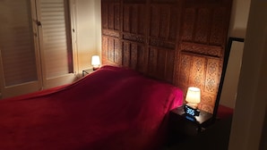 A hotel room with a bed covered in a red blanket, a nightstand with a lamp, and a wall with intricate wooden paneling. - Apartment F2, 4 pers, ideally located classified 2*, wifi  (Courseulles-sur-Mer)