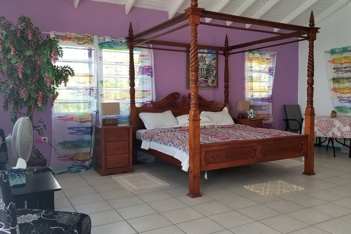 A four-poster bed with a wooden frame, a nightstand with a lamp, a chair, and a small table with a tablecloth.
