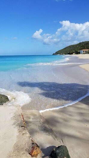 Une plage avec une eau turquoise et limpide, un rivage sablonneux et quelques bâtiments en arrière-plan.