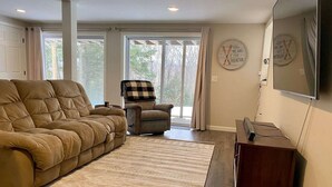 A living room with a large sofa, a smaller armchair, a coffee table, and a flat-screen TV. There are large windows with curtains, a rug, and a wall clock.