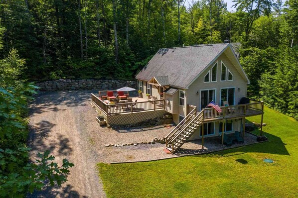 A two-story cabin with a deck, a stone wall, and a flag. - Gorgeous Retreat w/ 3 Spacious BRs & Decks, set on beautiful private acre! (Thornton)