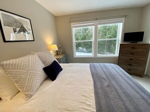 Une chambre à coucher comprenant un lit, une commode, un téléviseur et une fenêtre donnant sur des arbres.