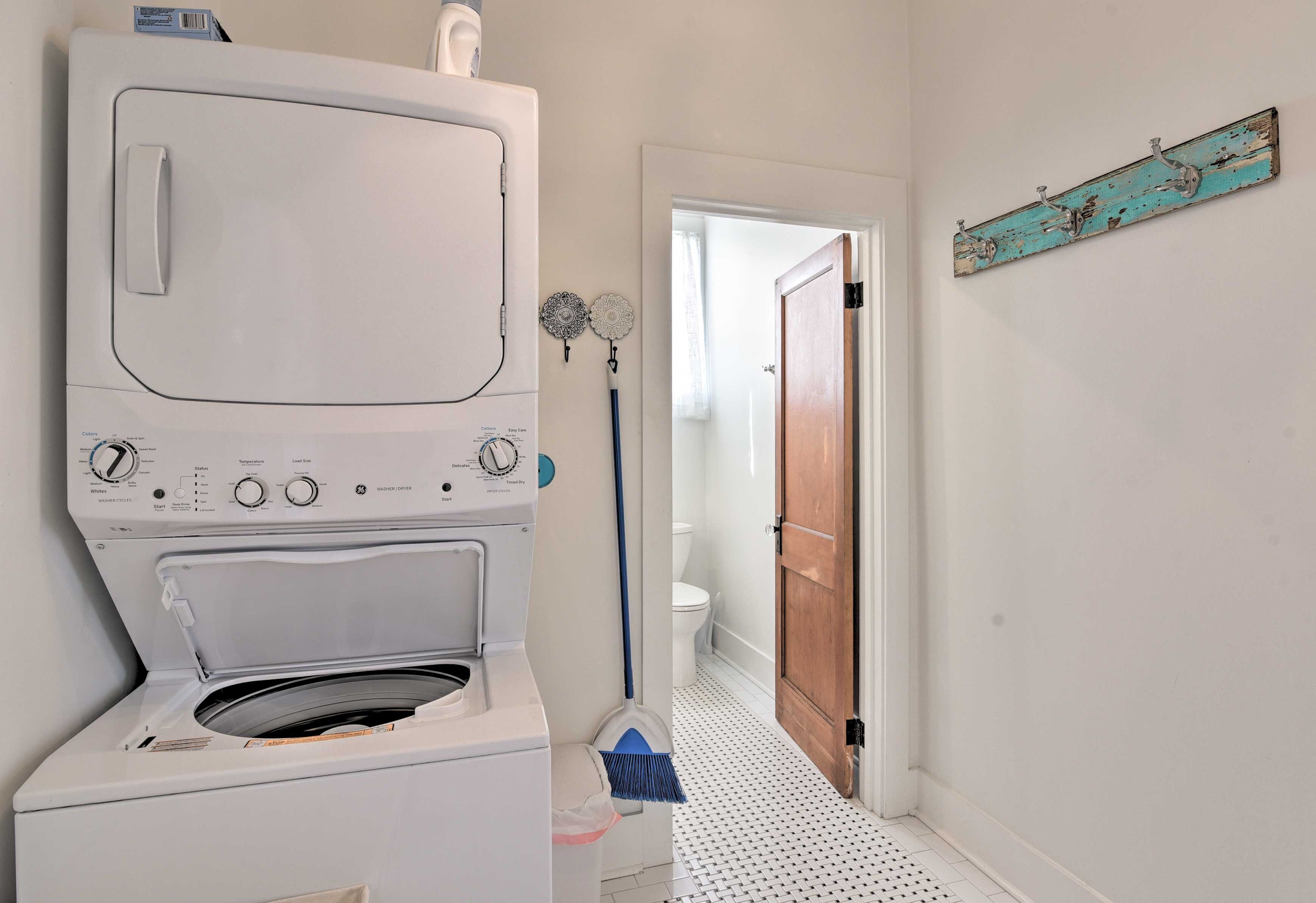A laundry room with a stacked washer and dryer, a broom, and a door leading to another room.