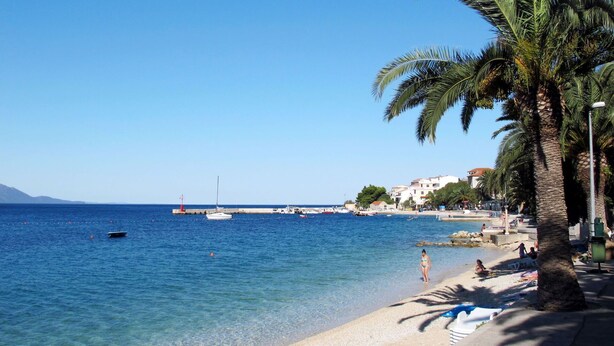 A beach with clear blue water, palm trees, and buildings in the background. - Cosy apartment for 4 guests with A/C, WIFI, TV, balcony and pets allowed (Gradac)