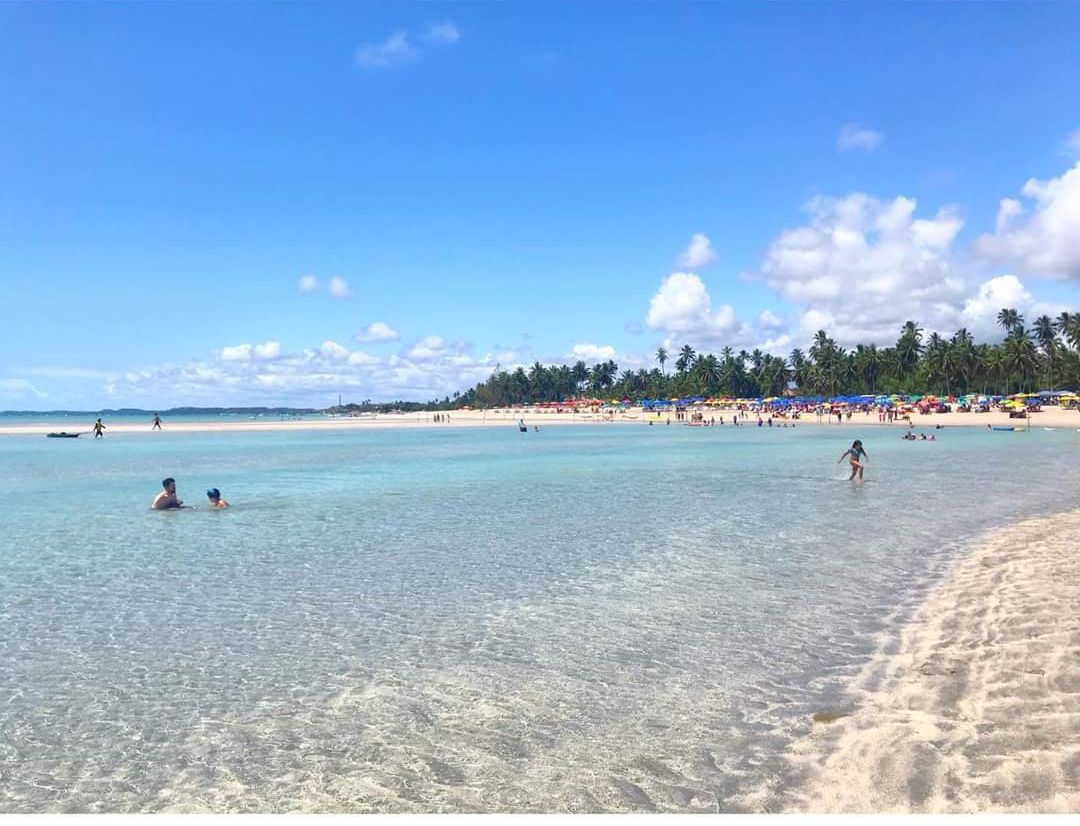 Una playa con aguas poco profundas y cristalinas, gente nadando y una orilla arenosa con sombrillas.