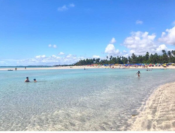 A beach with clear shallow waters, people swimming, and a sandy shore with umbrellas.