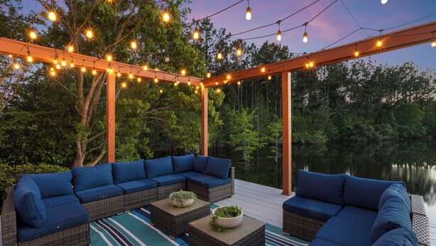 A covered outdoor seating area with wicker furniture and blue cushions, string lights, and a view of a lake. - Custom Built Lake House (Gulfport)