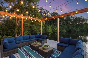 A covered outdoor seating area with wicker furniture and blue cushions, string lights, and a view of a lake.