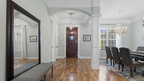 A hallway with a wooden floor, a bench, a large mirror, and a dining area with chairs and a chandelier. - Custom Built Lake House (Gulfport)
