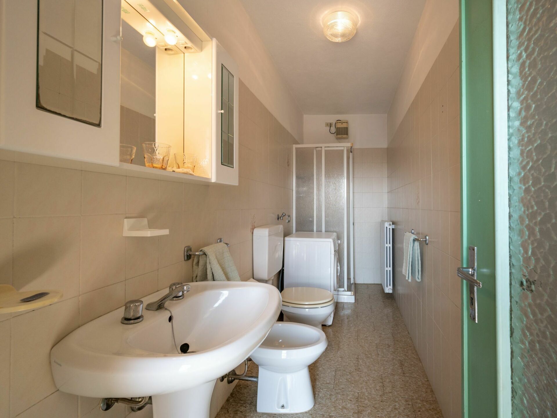A narrow bathroom with a sink, two toilets, and a shower stall.