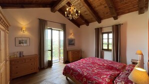 A bedroom with a wooden ceiling, a large bed with a red patterned bedspread, a chandelier, and a view through a large window. - Lovely apartment for 6 guests with pool, TV and terrace (Bonvicino)