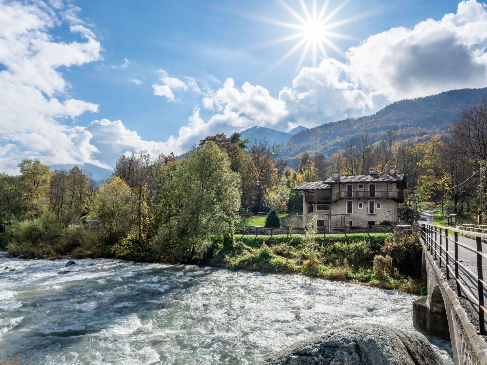 A stone house with a balcony overlooking a river, surrounded by trees and mountains.