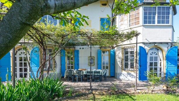 Un bâtiment doté d’un patio avec jardin, de volets bleus et d’un arbre au premier plan.