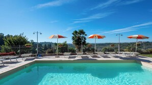A swimming pool with lounge chairs and umbrellas, surrounded by a fence and a view of the landscape.