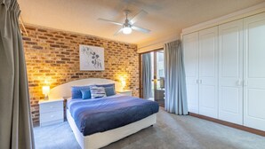 Une chambre avec un mur en briques, un ventilateur de plafond, un lit avec une literie bleue, une table de chevet avec une lampe et une porte-fenêtre coulissante donnant sur un espace extérieur.