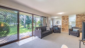 Un vaste espace de vie comprenant un canapé gris, un fauteuil plus petit et un meuble pour la télévision. Une porte-fenêtre coulissante donne accès à un jardin.