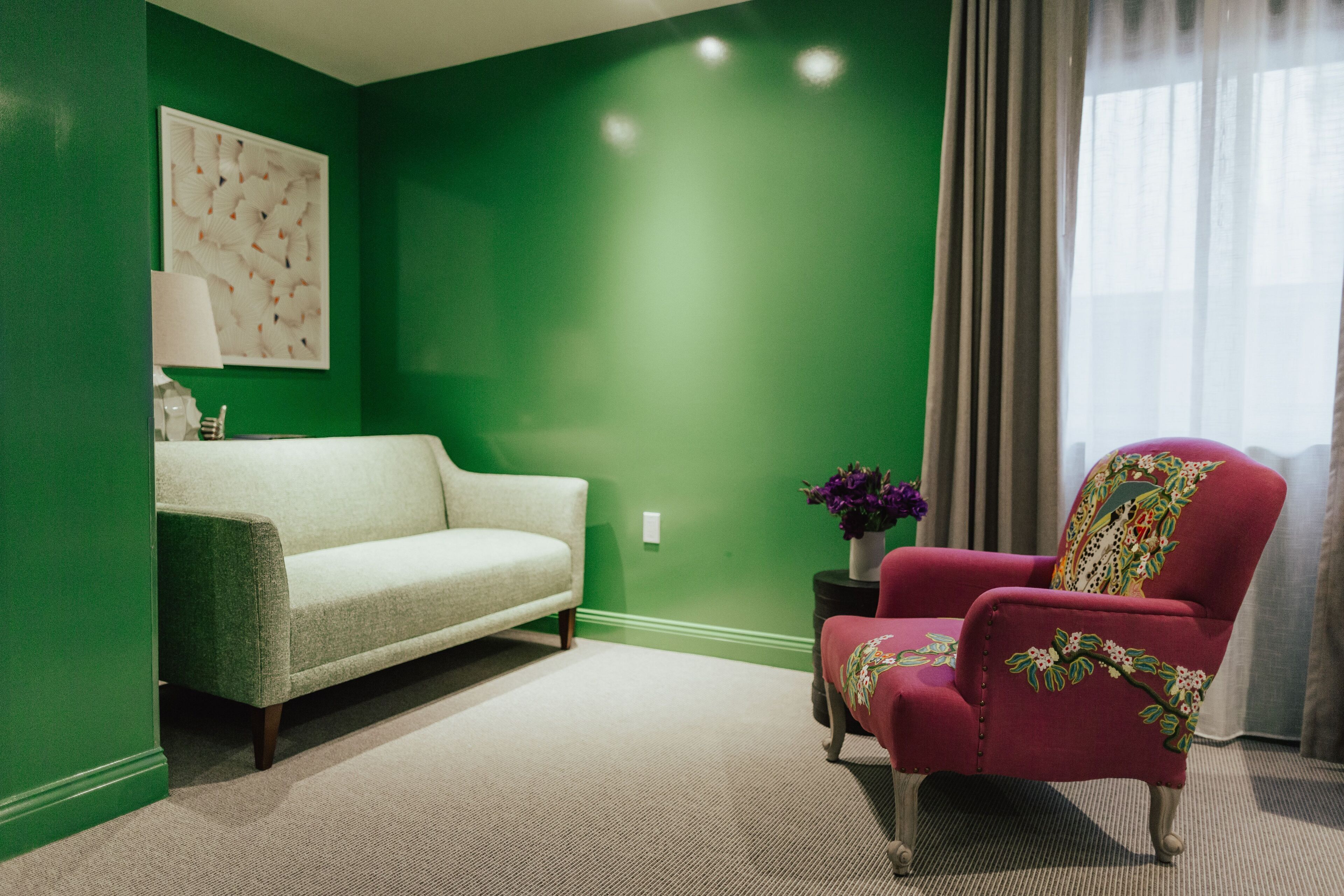 A room with a green wall, a white sofa, a red armchair, a small side table with a vase of flowers, and a window with curtains.